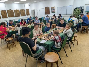 Adults and juniors playing a chess tournament together in the Hammersmith Chess Club main hall, with wooden chess boards and sets. Mentors are sitting next to each set of players. There are about 25 people in the room.