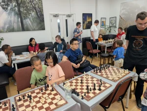 A mix of adults and children sit and stand around several chess boards, reviewing and discussing chess games which have just happened, using the boards for analysis.