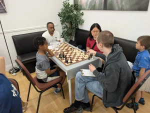 Two men, a woman and two boys sit around a wooden chess board, discussing a game that has just happened. One of the adults is moving the pieces around to explain the game to the others.