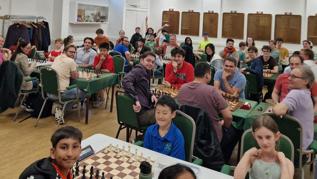 A room full of colourfully dressed chess players, seated at their chess boards, look and wave excitedly at the camera