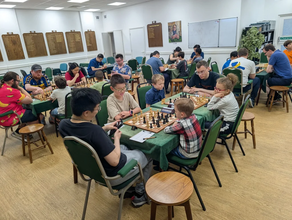 Adults and juniors playing a chess tournament together in the Hammersmith Chess Club main hall, with wooden chess boards and sets. Mentors are sitting next to each set of players. There are about 25 people in the room.