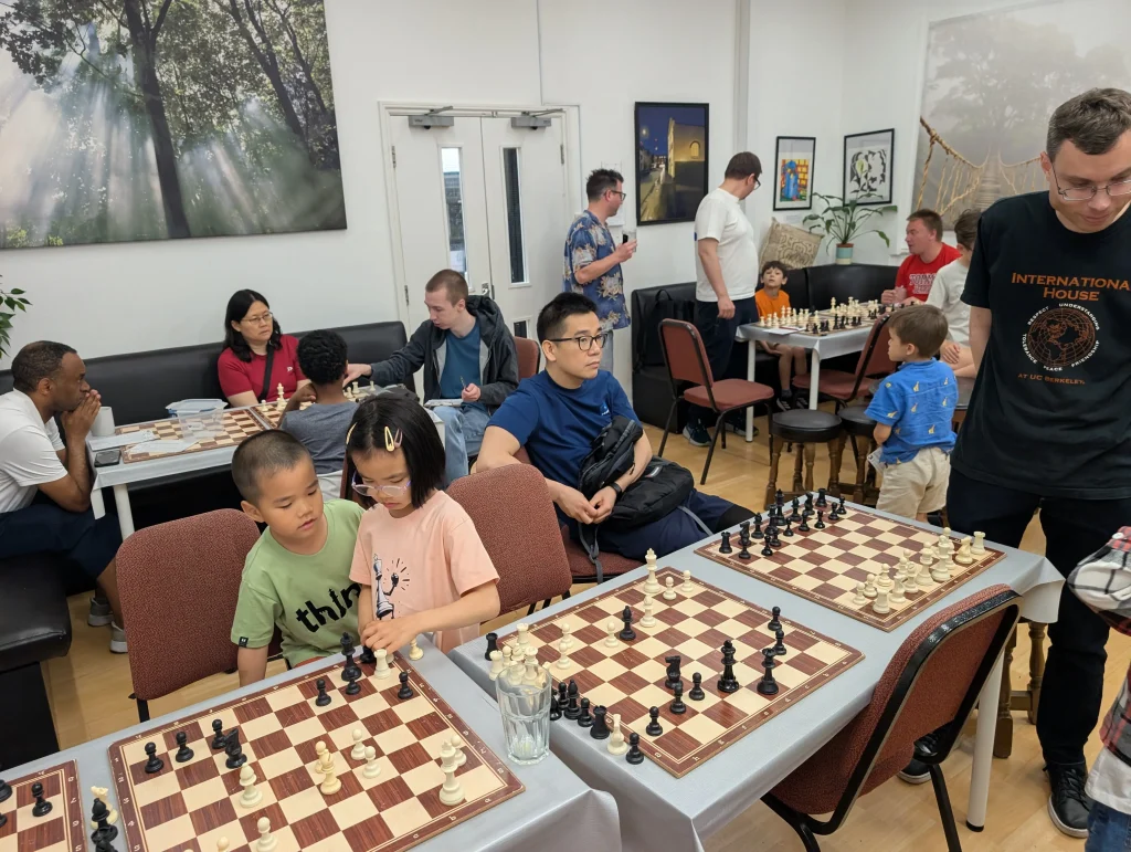 A mix of adults and children sit and stand around several chess boards, reviewing and discussing chess games which have just happened, using the boards for analysis.