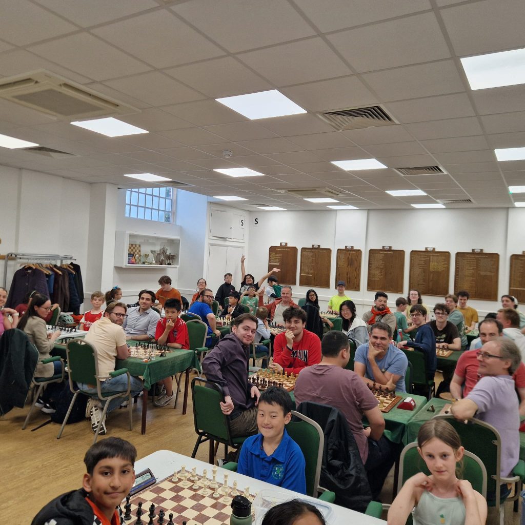 A room full of colourfully dressed and excited chess players, kids and adults, sitting at their boards and looking or waving at the camera.